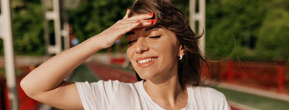 Mulher branca e de cabelos castanhos protegendo o rosto do sol para evitar o envelhecimento cutaneo Fotoproteção