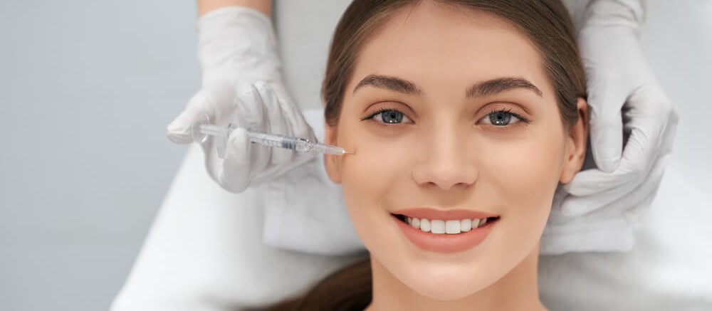 Moça de cabelos castanhos sorridente aplicando uma injeção para rejuvenescimento facial